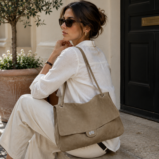 A woman in a white linen outfit and sunglasses sits on stone steps beside a black door, carrying a taupe suede shoulder bag with a flap closure.