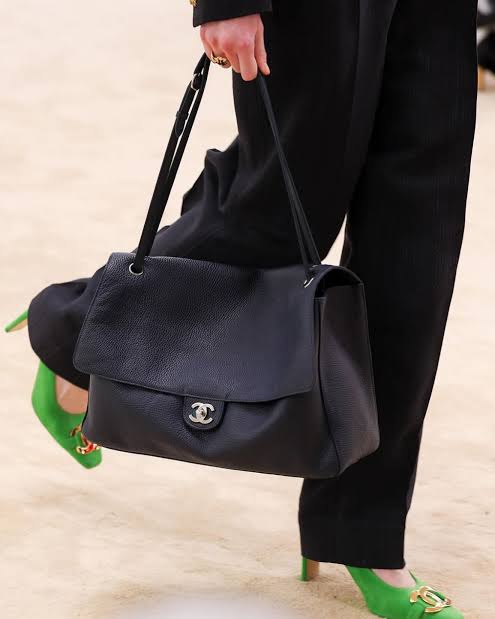 Person holding a black Chanel handbag with green shoes on a light background