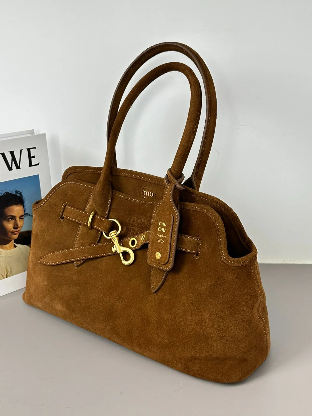 Brown suede handbag with gold hardware on a light gray surface.