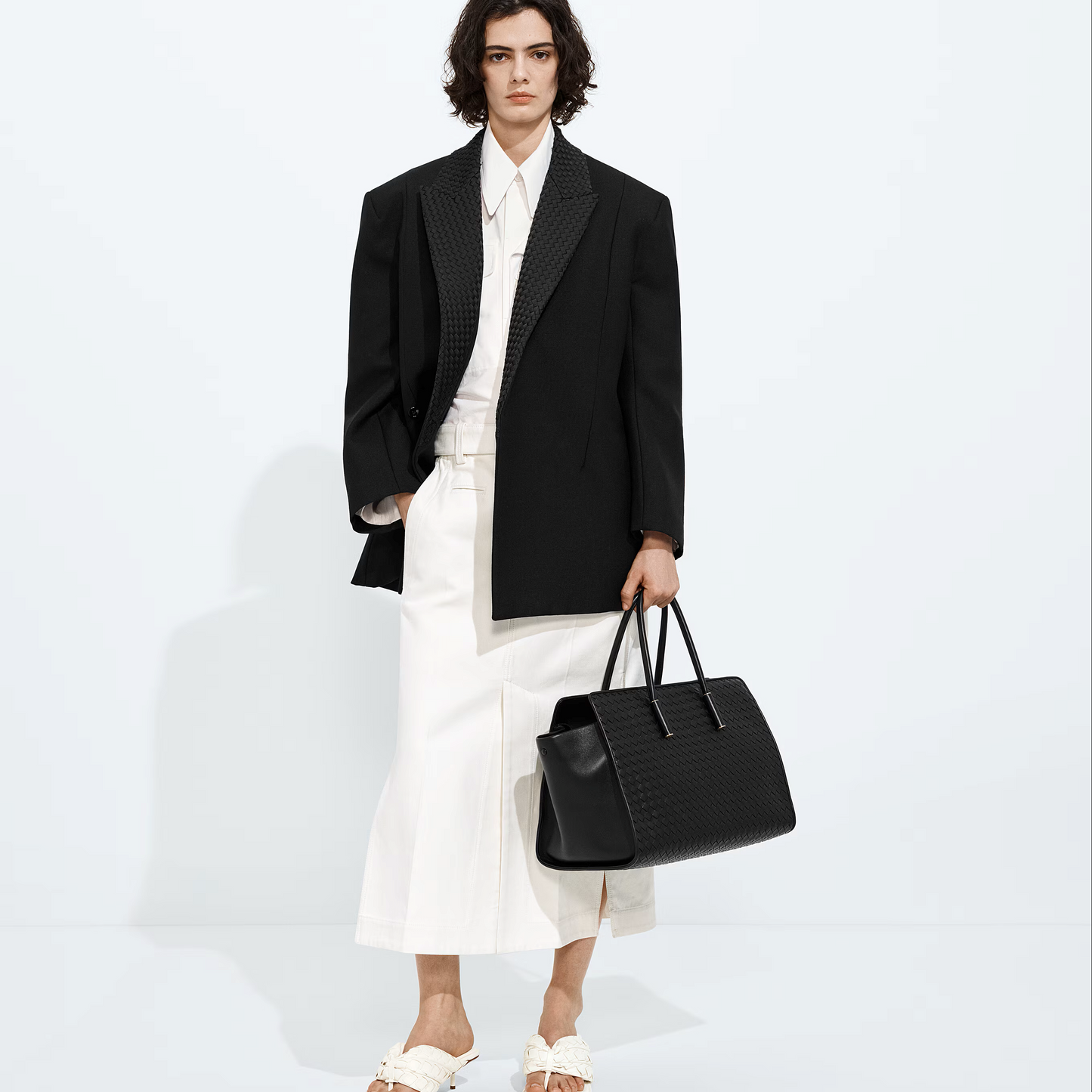 A model stands against a clean white studio background wearing a black textured blazer, white shirt, white pleated skirt, and white slide sandals, while holding a black crocodile-embossed leather tote bag.
