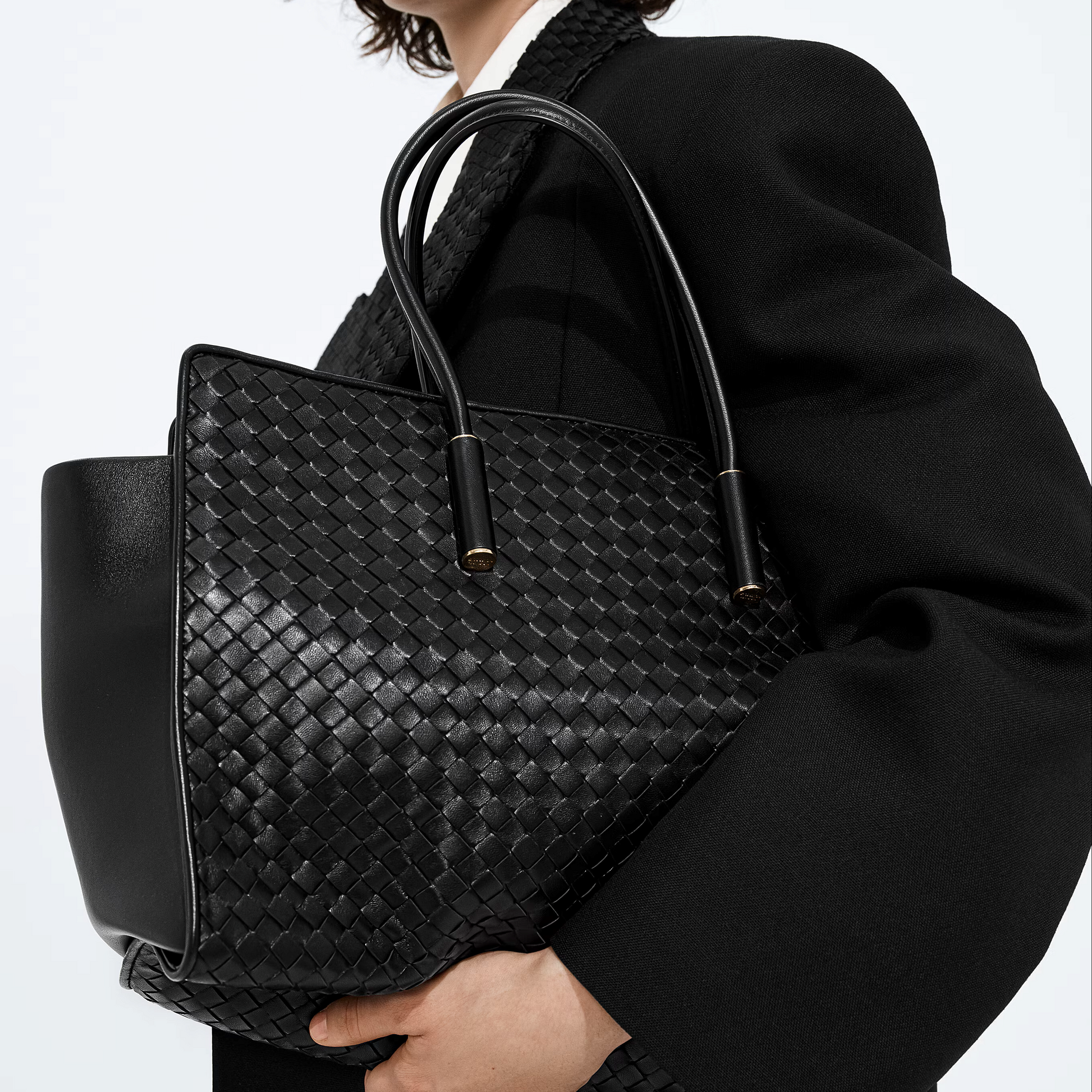 A person in a black coat holds a large black woven leather tote bag with double top handles against a plain light background.
