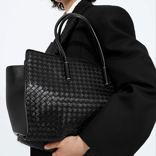 A person in a black coat holds a large black woven leather tote bag with double top handles against a plain light background.