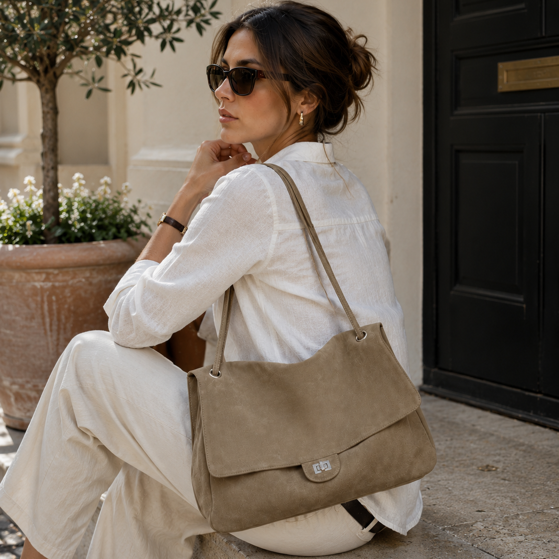 A woman in a white linen outfit and sunglasses sits on stone steps beside a black door, carrying a taupe suede shoulder bag with a flap closure.